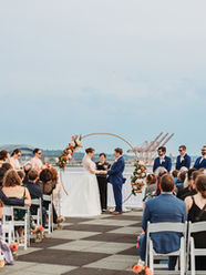 A photo of a wedding ceremony in progress with the bride and groom holding hands at the altar. They are standing in front of a circular gold arch with pink and orange flower installations on either side. Behind them is the south Seattle skyline. Their guests are sitting in front of them in white chairs while their bridal party stands on either side of them wearing sunglasses.