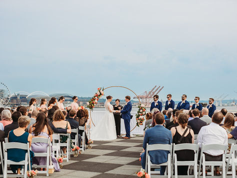 A photo of a wedding ceremony in progress with the bride and groom holding hands at the altar. They are standing in front of a circular gold arch with pink and orange flower installations on either side. Behind them is the south Seattle skyline. Their guests are sitting in front of them in white chairs while their bridal party stands on either side of them wearing sunglasses.