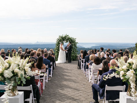 Photo of a wedding ceremony with the bride and groom at the altar holding hands while their guests watch. The couple is framed by a lush arch made from greenery and white flowers. Behind them is the Seattle skyline on a partly cloudy day.