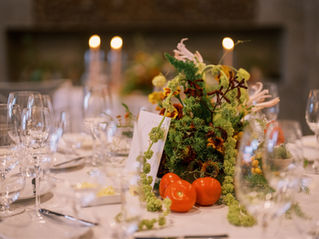 Close up photo of a wedding reception table with white plates and simple glassware and flatware. The centerpiece is made up of flowers, greenery, and tomatoes!