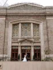 A bride and groom on their wedding day standing in front of The Sanctuary at Lotte Hotel Seattle, an over 100 year old former church made of bricks and stained glass. They are standing in front of the doors looking at the camera.