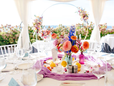 Close up photo of a wedding reception table. The centerpiece is made up of a few flower arrangements in different colored glass vases and the flowers are in bold, vibrant colors. There are lemons and blooms on the table as well. The centerpiece is surrounded by a muted purple guaze runner that is in a circle around the decor. Behind the table is the sweetheart table with a large floral arch and in the background are more flower bushes.