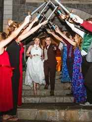 Photo of a bride and groom's grand exit from their wedding. They are walking down a stone staircase and their guests are lining both sides of the stairs, raising swords over their heads as they walk out.
