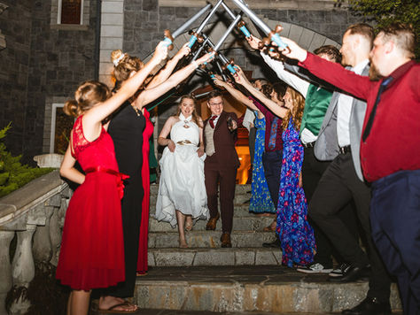 Photo of a bride and groom's grand exit from their wedding. They are walking down a stone staircase and their guests are lining both sides of the stairs, raising swords over their heads as they walk out.