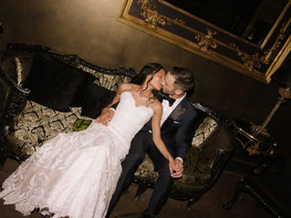Photo of a bride and groom sitting on an ornate, vintage couch at their wedding. They are holding hands and kissing. The room is dark and they are being lit by the camera flash.