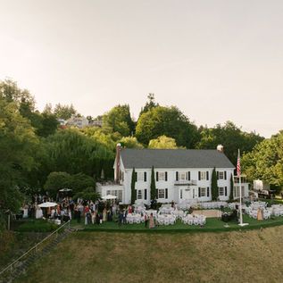 Drone photo of The Admiral's House set up for a wedding. There are reception tables with a dance floor in the center in the back of the  house. Off to the side of the house, there are guests mingling for cocktail hour under umbrellas. The house is framed by lush green trees and the house is on a cliffside that is partially visible in the photo. 