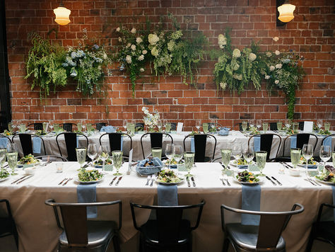 Photo of a wedding reception dinner tables. The tables are long with white linens and blue napkins draped off the edge. Each place setting has a salad with bread, water, and wine glasses also on the table. The chairs are modern, and grey metal style. The backdrop is a red brick wall with lush floral installations hanging on the wall. there are light fixtures hanging from the ceiling as well. 