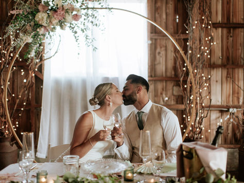Bride and groom share a kiss at their sweetheart table, celebrating with champagne beneath a twinkling arch