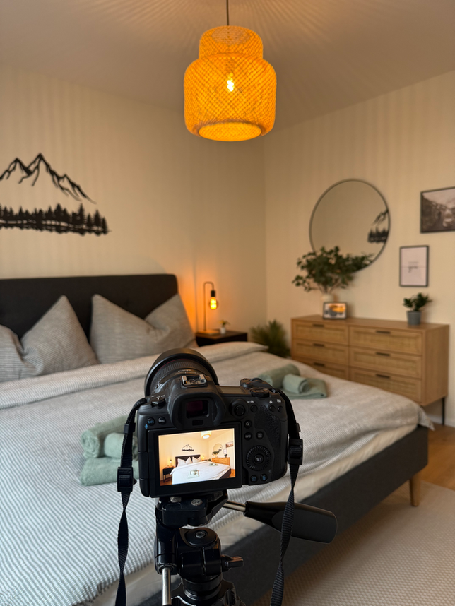 Im Vordergrund sieht man eine Kamera auf einem Stativ. Dahinter sieht man ein Schlafzimmer mit einem Doppelbett, Pflanzen und Nachttischen mit Lampen in einer Ferienwohnung in Spiez, Bern.
