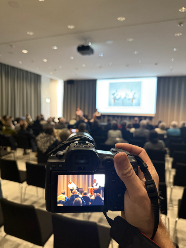 Im Vordergrund sieht man eine Hand die eine Kamera hält. Dahinter sieht man einen Saal, eine grosse Leinwand, zwei Personen die sitzen zu einem Publikum in Lausanne sprechen.
