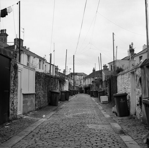 Eine etwas heruntergekommene Kopfsteinpflaster Hintergasse mit zahlreichen Abfallcontainern im St. Judes Quartier, Plymouth, England, schwarz weiss Foto.