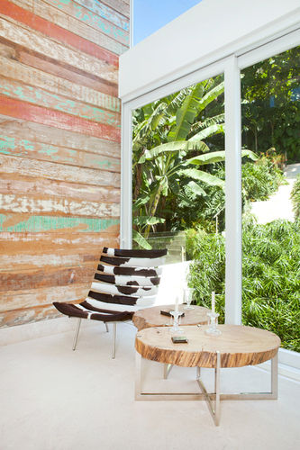 Detailansicht eines Wohnbereichs mit einem Sessel und zwei Tischen vor einer Holzwand und einem Fenster mit Aussicht auf einen begrünten Garten in Joá, Rio de Janeiro, Brasilien.