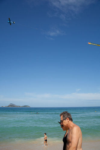 Ein Paar am Strand von Leblon, ein Junge und Surfer im Meer, im Hintergrund die die Ilhas Cagarras und ein Flugzeug am Himmel mit einem Werbebanner in Rio de Janeiro, Brasilien.