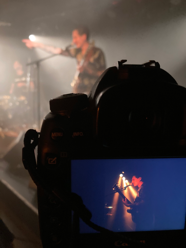 Im Vordergrund die schwarze Silhouette einer Kamera mit der Bildvorschau einer Konzertfotografie von Mathias Schenk. Im Hintergrund unscharf die Band Hello Cleveland beim Spielen eines Konzertes im Le Singe in Biel.