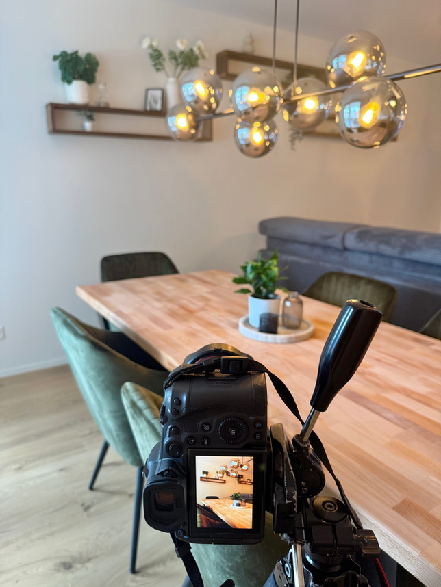 Im Vordergrund sieht man eine Kamera auf einem Stativ. Im Hintergrund sieht man ein Wohnzimmer mit einem Tisch, Stühlen und einer Design Lampe in einer Wohnung in Spiez.