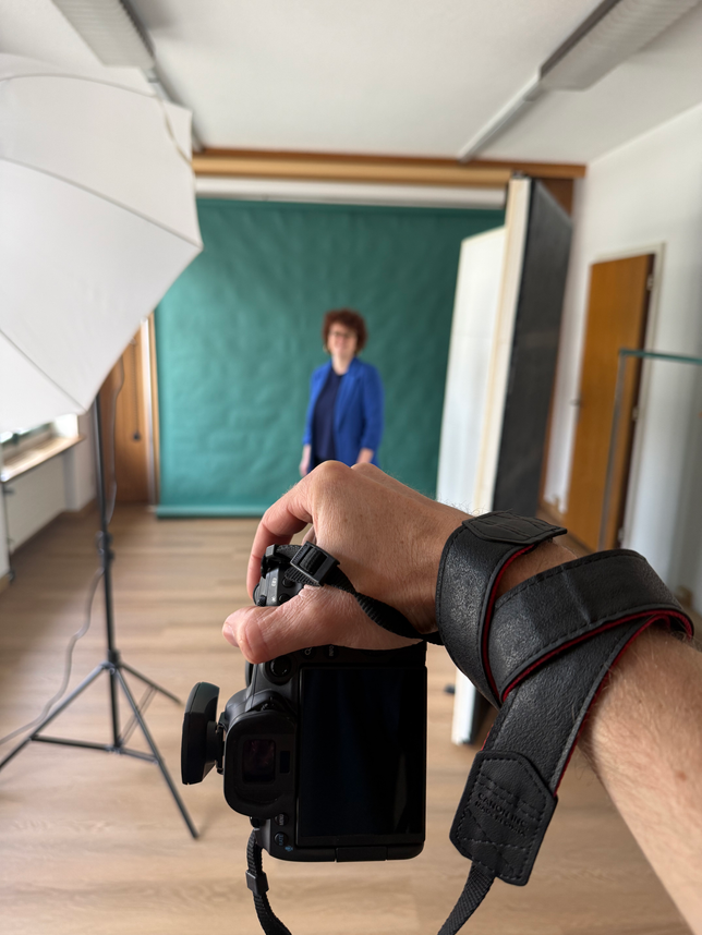 Im Vordergrund sieht man eine Hand die eine Kamera mit Blitzauslöser hält. Die Kamera ist auf eine Frau vor dunkelgrünen Hintergrund gerichtet. Sie ist umgeben von Reflektoren und Blitzlampen im Fotostudio in Biel.