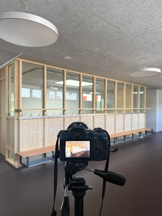 Im Vordergrund sieht man eine Kamera auf einem Stativ. Dahinter sieht man einen Teil eines Kindergartens in Biel mit einem Garterobenbereich aus Holz und Glas.