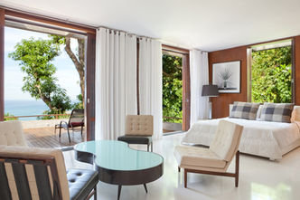 Ein grosses Schlafzimmer mit vier Sesseln, einem Tisch und Bett mit einer grossen Terrasse und Blick auf den Wald und das Meer in São Conrado, Rio de Janeiro, Brasilien