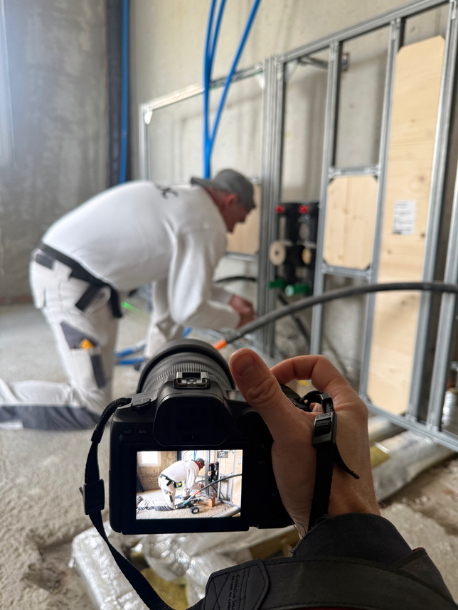 Im Vordergrund sieht man eine Hand die eine Kamera hält. Die Kamera ist auf einen Arbeiter in weisser Kleidung auf einer Baustelle in Lengnau gerichtet.