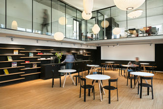 Café Bereich der Bibliotheken Köniz mit drei Personen mit Bewegungsunschärfe. Im oberen Teil sieht man die Büroräunlichkeiten der Bibliothek in Köniz bei Bern.