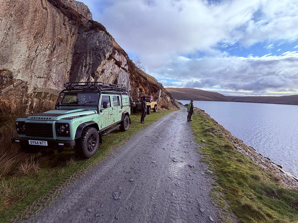 Two Land Rover Defenders parked beside a narrow reservoir track in the Elan Valley, with steep cliffs and open water creating a scenic mid-journey stop.