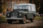 Classic Land Rover Series III with green body and white roof, parked on a grassy area with trees and buildings in the background.