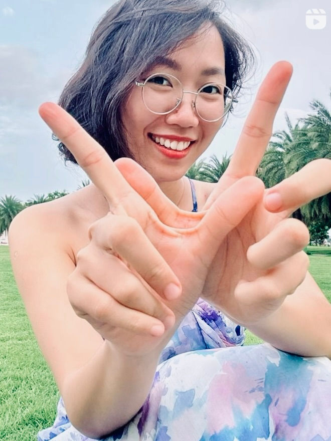 Woman smiling, forming a hand gesture, wearing glasses and a colorful dress. Green park with palm trees in the background, bright mood.