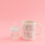 A baby bottle and a white mug with "CHEERS Y'ALL" text on a pink background. Contrast of infant and adult themes. Playful mood.