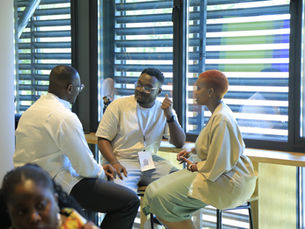 Three young professionals sitting around a high table engaged in a discussion. The man in the center, wearing a beige t-shirt and glasses, is speaking and gesturing with his hand to the woman on the right, who has short orange-red hair. The third man on the left is listening. The background shows large windows with horizontal blinds.