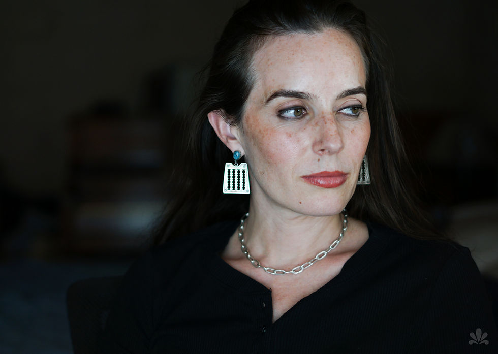 Woman with long hair gazes sideways, wearing black top, silver chain necklace, and unique rectangular earrings.