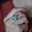 Thumbnail: Hand with silver ring featuring an oval turquoise gemstone. The background is softly blurred, worn on a finger