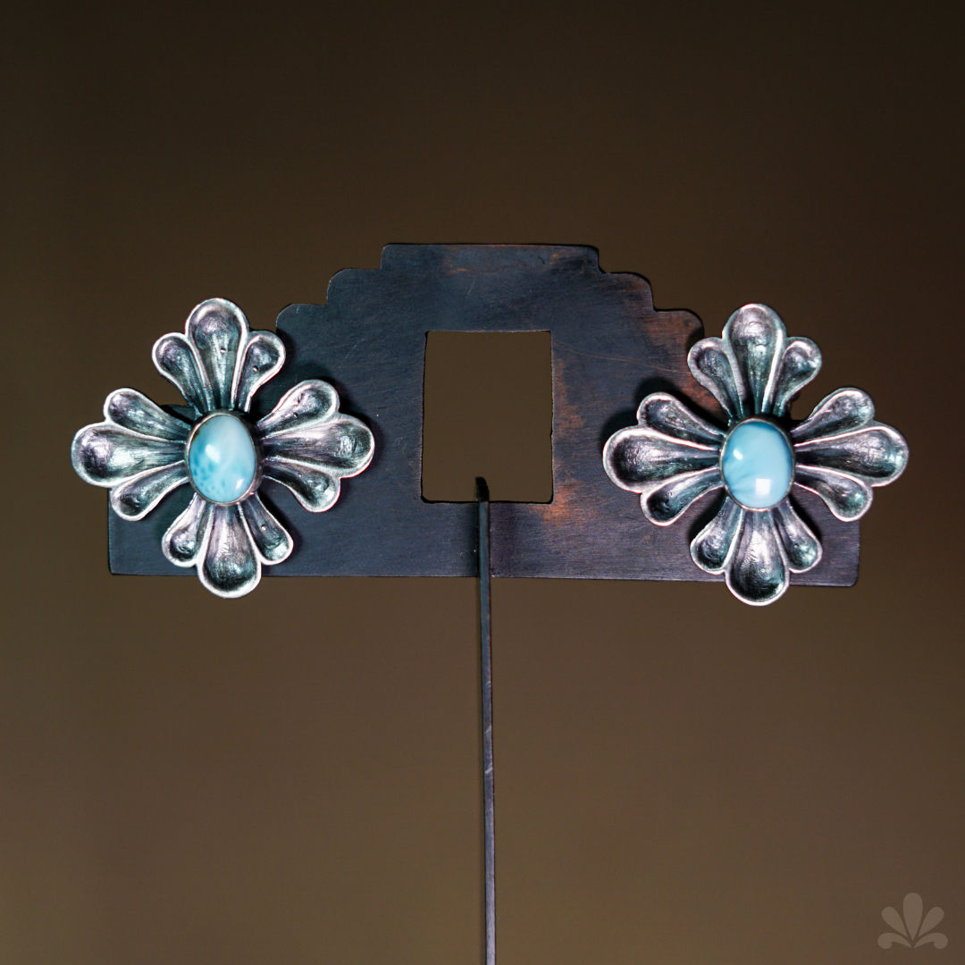 Two silver floral earrings with blue stones displayed on a dark stand against a brown background, giving an elegant vibe