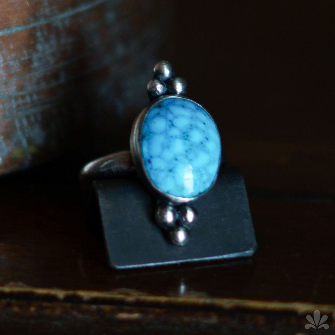 Silver ring with an oval blue stone featuring black speckles, set on a black stand against a wooden background