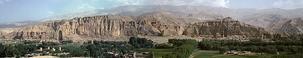 Panoramic view of Bamiyan cliffs with lush green fields below. Blue sky and mountains in the background, evoking a serene landscape.