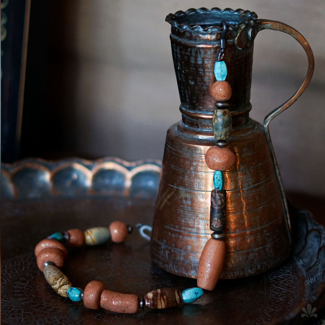 Beaded necklace draped over copper vase. Background has a textured, speckled pattern. Earthy tones.