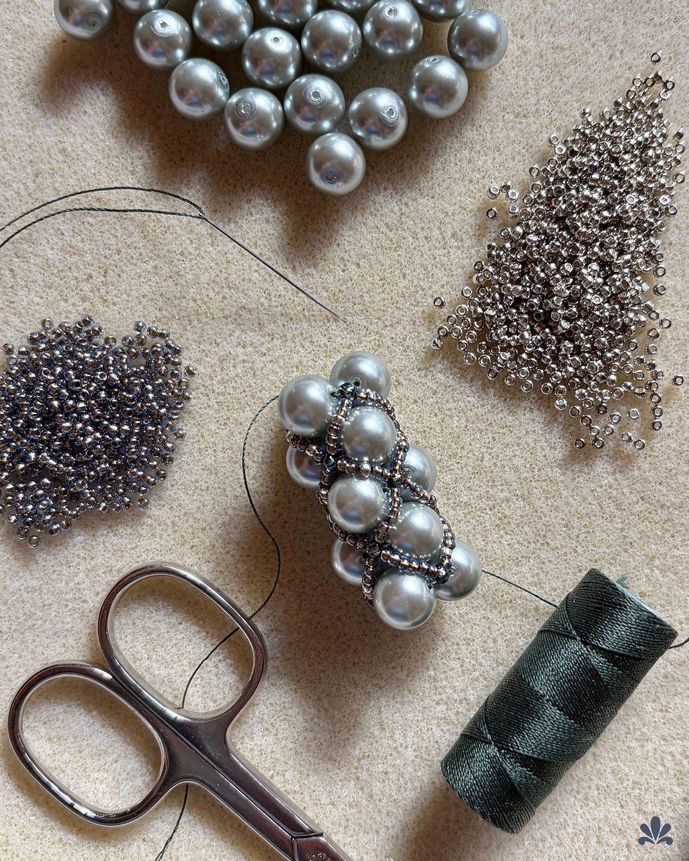 Beads and pearls on fabric, with silver scissors and green thread. A needle threads through pearls, surrounded by silver beads. Calm, creative scene.