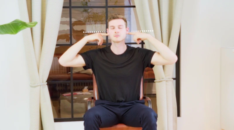 Person sitzt auf einem Stuhl und hält seine Zeigefinger an die obere Halswirbelsäule und hat die Augen dabei geschlossen. 