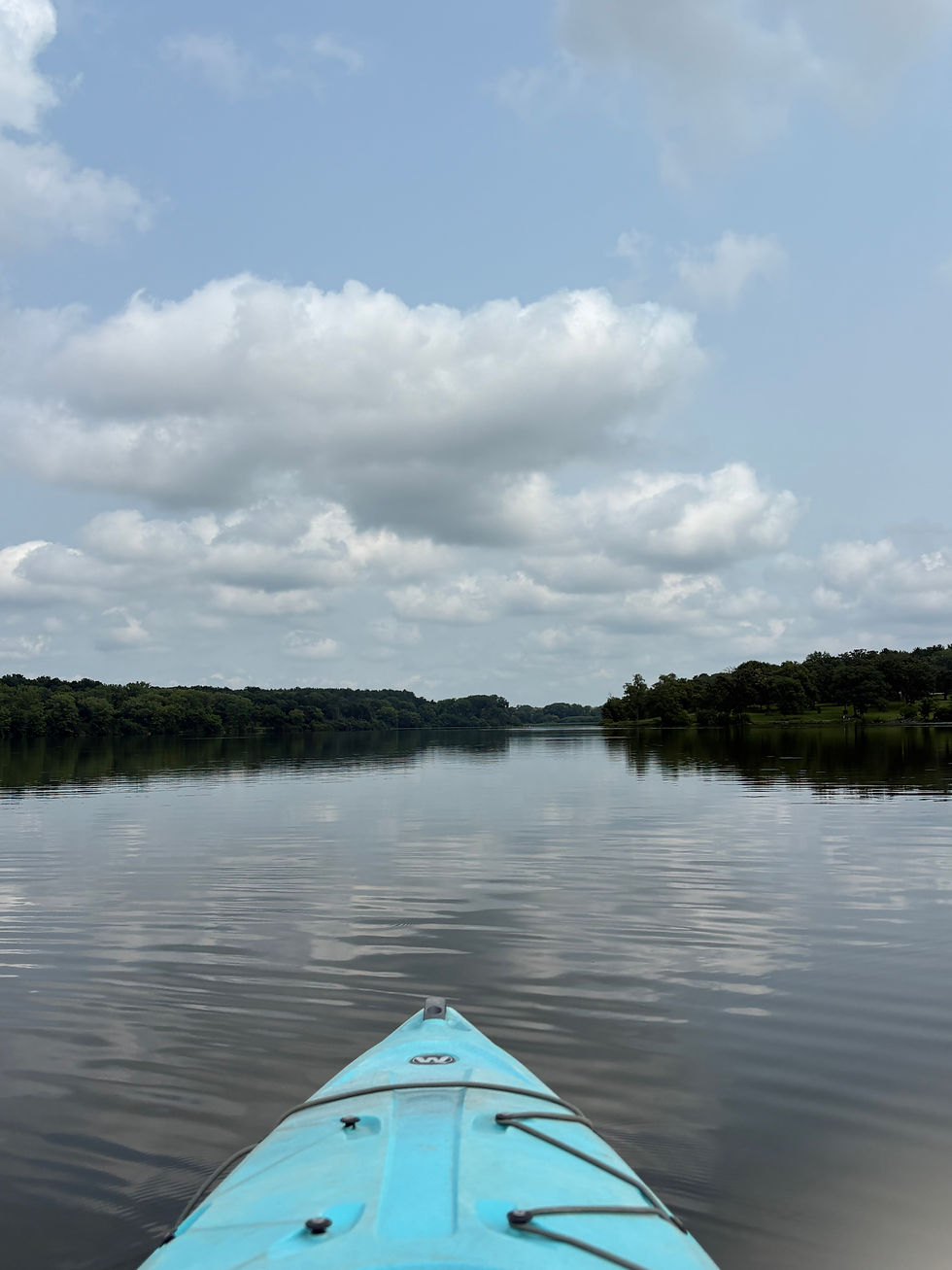 Blue kayak bow on a calm lake, with cloudy sky and tree-lined shores reflected in the water.