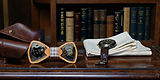 Handmade wooden bowties and a watch on a wooden surface with books in the background.
