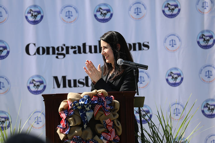 A person stands at a podium, clapping, with a banner that reads "Congratulations Mustangs" in the background.