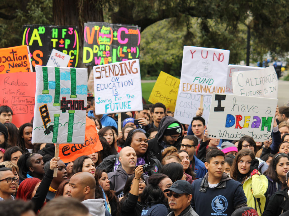 Crowd protests, holding signs: "SPENDING FOR EDUCATION FUTURE", "FUND EDUCATION" outdoors.