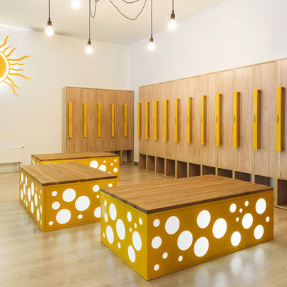 Návrh interiéru šatny pro mateřskou školu s důrazem na moderní design, dřevěné prvky, žluté lavice a osvětlení.
