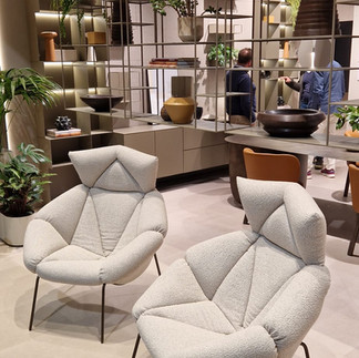 Designová zóna na veletrhu Salone del Mobile 2025 s moderními křesly a otevřenou kovovou knihovnou s dekoracemi a zelení.