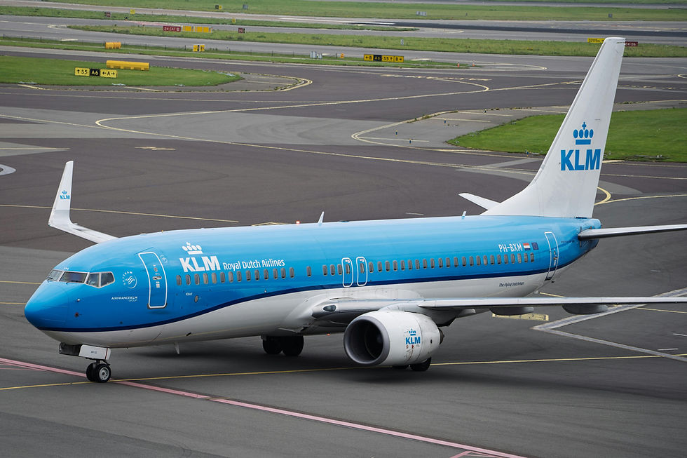 KLM airplane with blue and white livery taxis on runway. KLM logo visible on tail and fuselage. Overcast sky and airport signs in background.