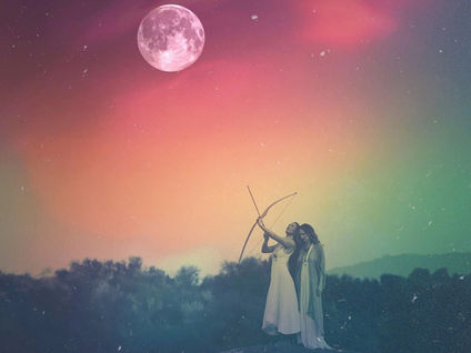 Two women in flowing white dresses standing on a rock under a luminous pink full moon, one drawing a bow and aiming at the sky against a dreamy rainbow-colored night sky, representing the Sagittarius Full Moon invitation to aim for something larger than yourself and trust the divine with the rest