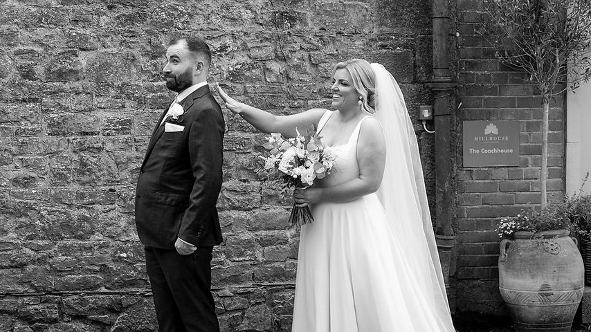 A bride standing behind her husband groom is tapping him on the shoulder. Both are wearing wedding apparel; he is wearing a suit and she is wearing a white wedding dress and a veil, holding a wedding bouquet. A brick wall is behind them. The couple is outside. 