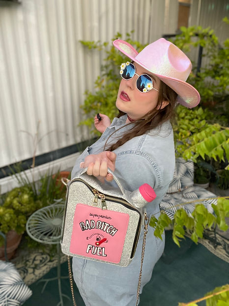 Plus size woman in denim jumpsuit and a pink cowboy hat poses with a purse that says "bad bitch fuel." She looks sassy.