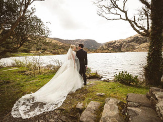 a bride with a long wedding dress stands beside her new husband beside a lake