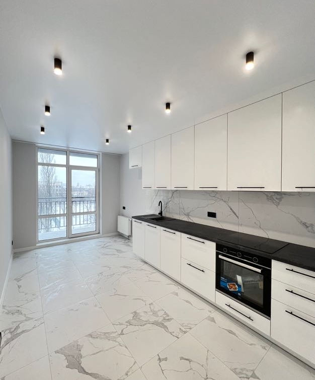 Сучасна кухня з білими шафами, чорною стільницею, великим вікном та освітленням.