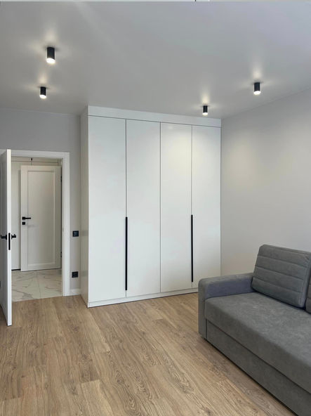 Біла шафа в кімнаті з сірим диваном, двері відчинені. ПРОЕКТИ Key4Comfort